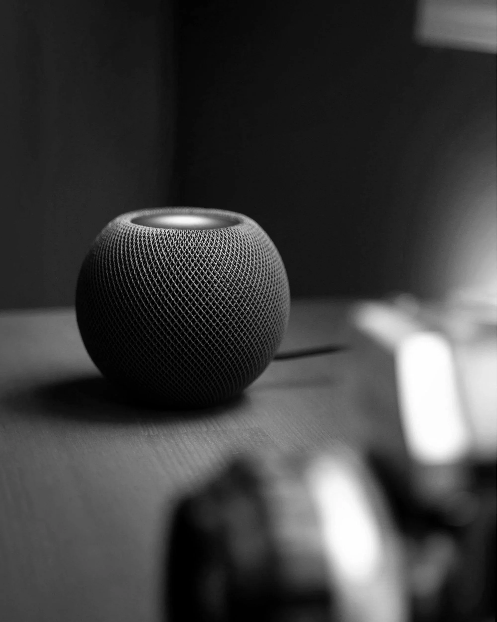A close-up, black-and-white image of a small speaker or smart device with a textured surface, placed on a smooth surface with blurred background.