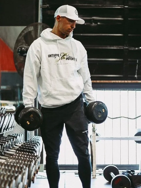 A man in a white hoodie and black pants lifting dumbbells in a gym.