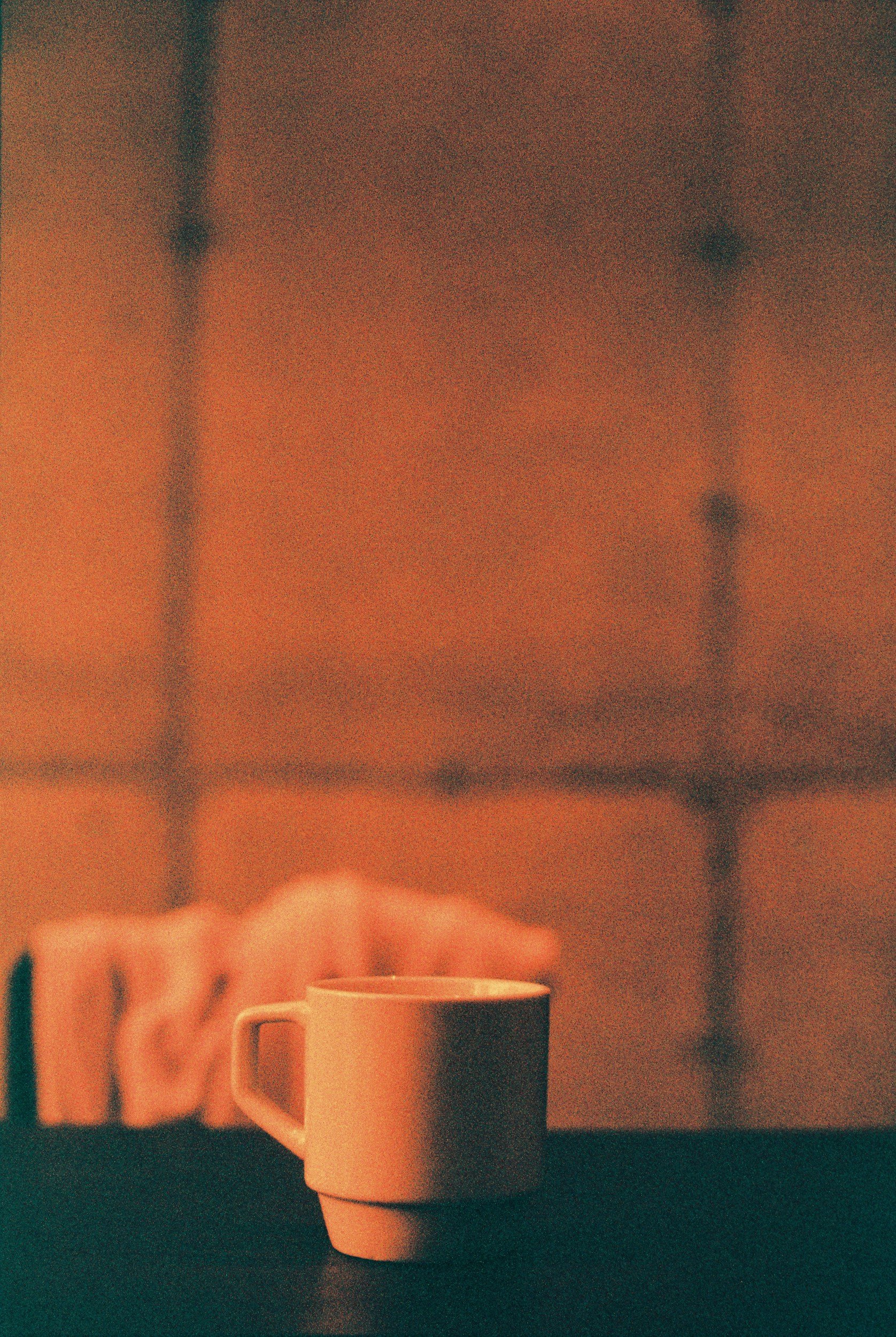 A beige ceramic coffee mug on a dark surface with a background of blurred, orange-colored wall tiles and black grout lines.