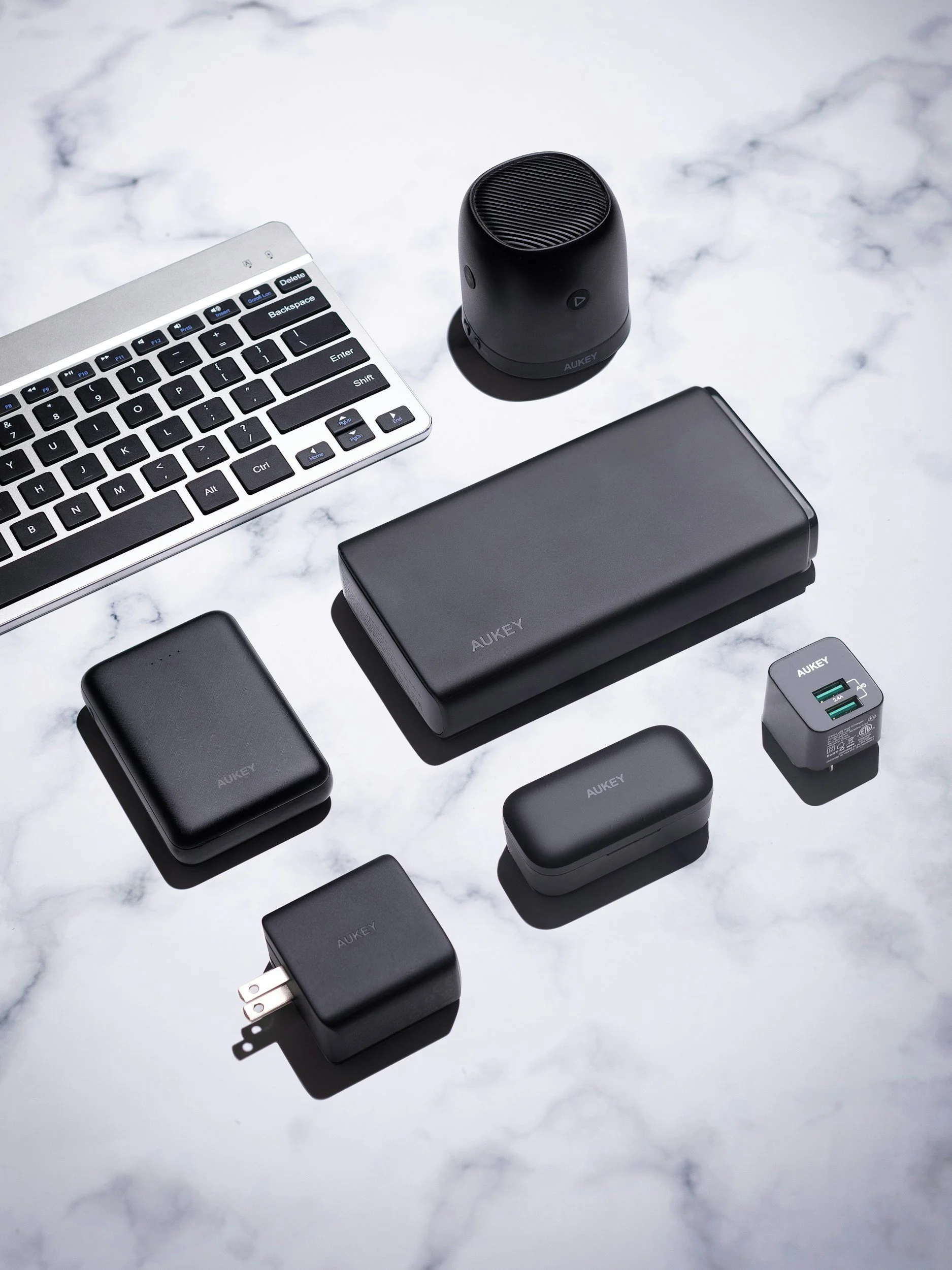 Collection of electronic accessories including a wireless speaker, portable power banks, a USB wall charger, and other small gadgets on a marble surface.