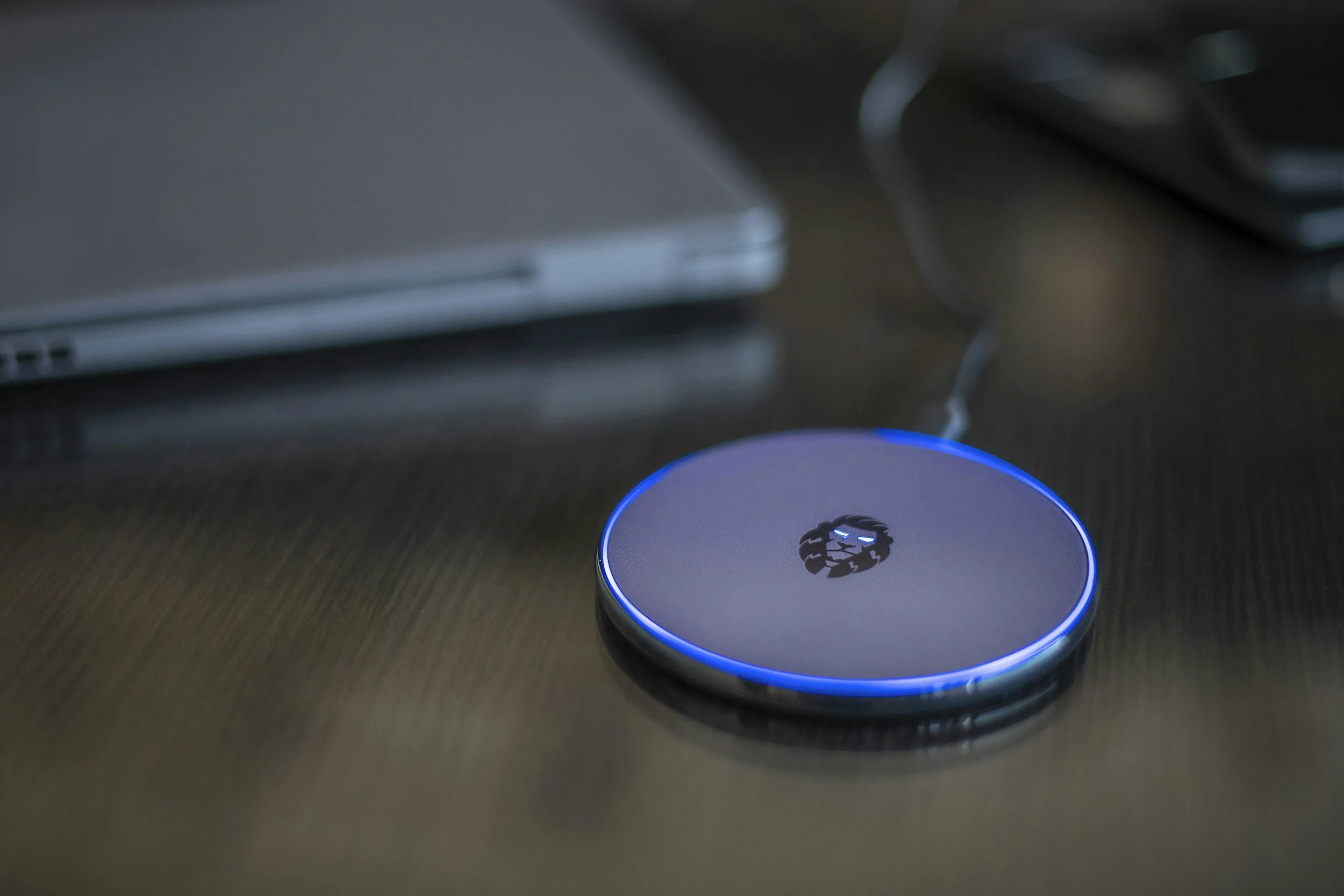 A circular electronic device with a lion logo in the center and a blue LED light around the edge, placed on a wooden surface near a closed laptop and a black object.