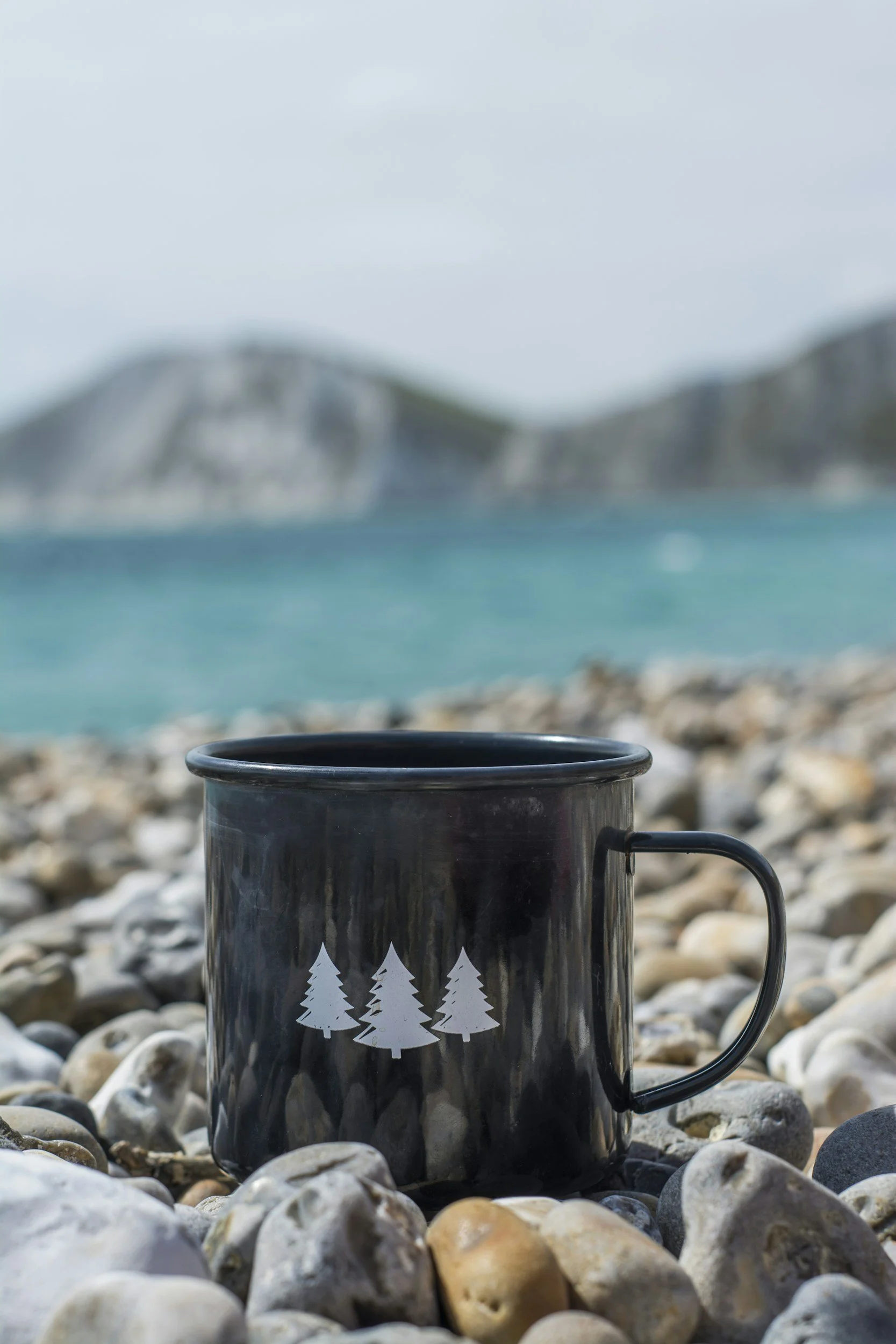 A black metal mug with white Christmas tree designs, placed on a rocky beach with the ocean and distant cliffs in the background.