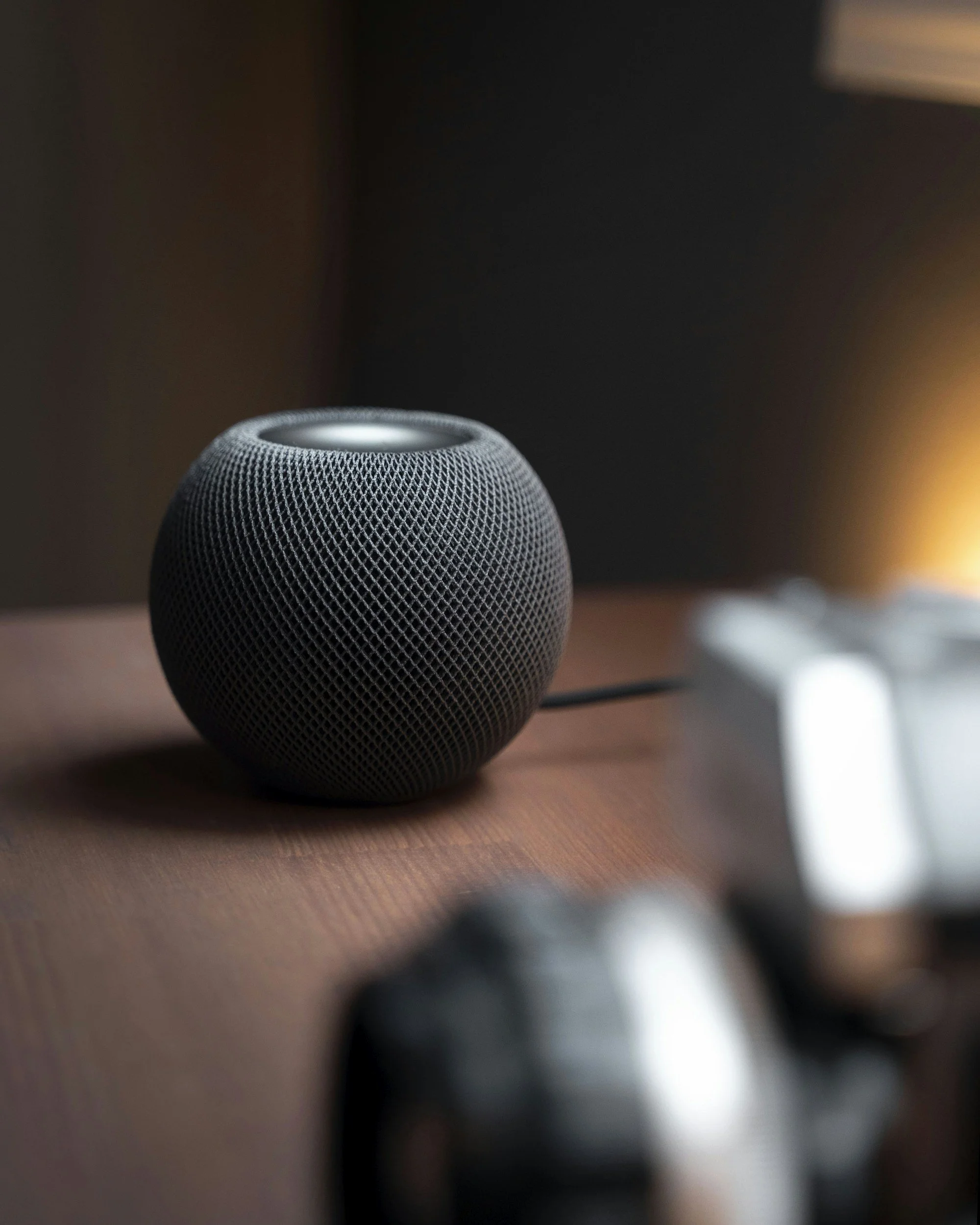 A close-up of a smart speaker with a mesh exterior, placed on a wooden surface with a blurred warm light in the background.