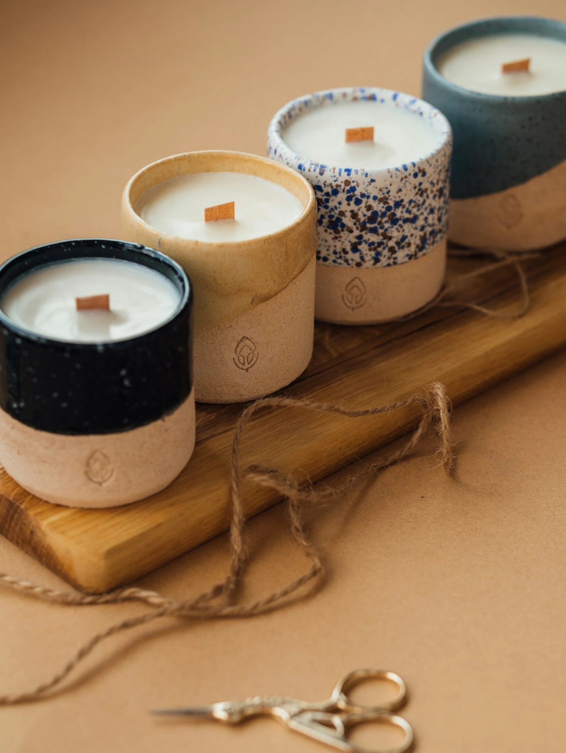 Four candles in ceramic holders with unique speckled and solid-colored designs, placed on a wooden board with scissors and a piece of string nearby.