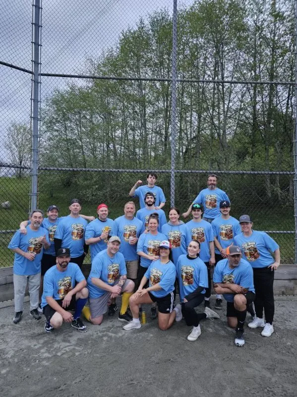 Group of people in matching blue T-shirts posing on a baseball field near a tall chain-link fence with trees in the background.