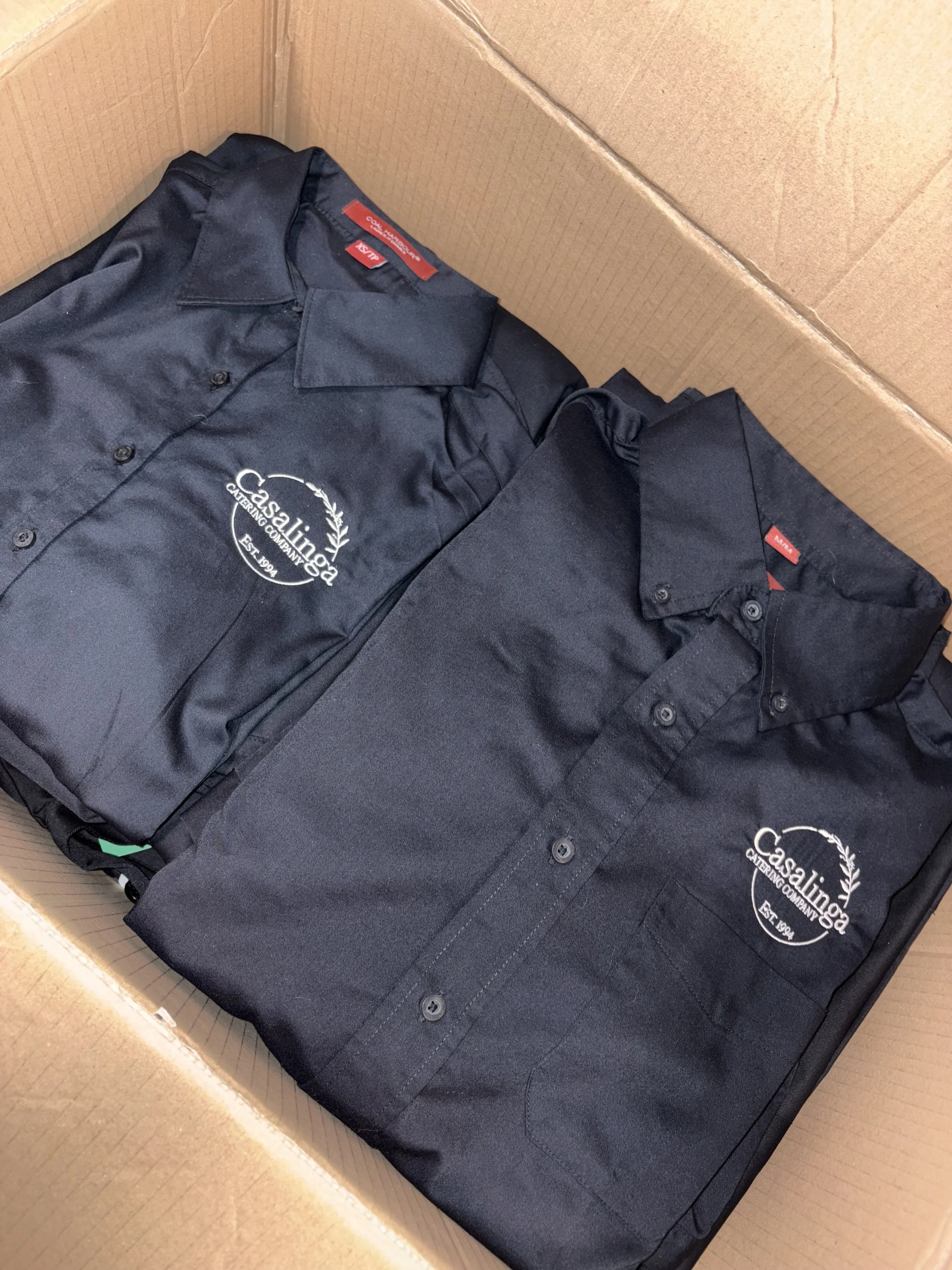 A cardboard box containing black shirts with a white logo that reads 'Casalinga Catering Company Est 1994.'