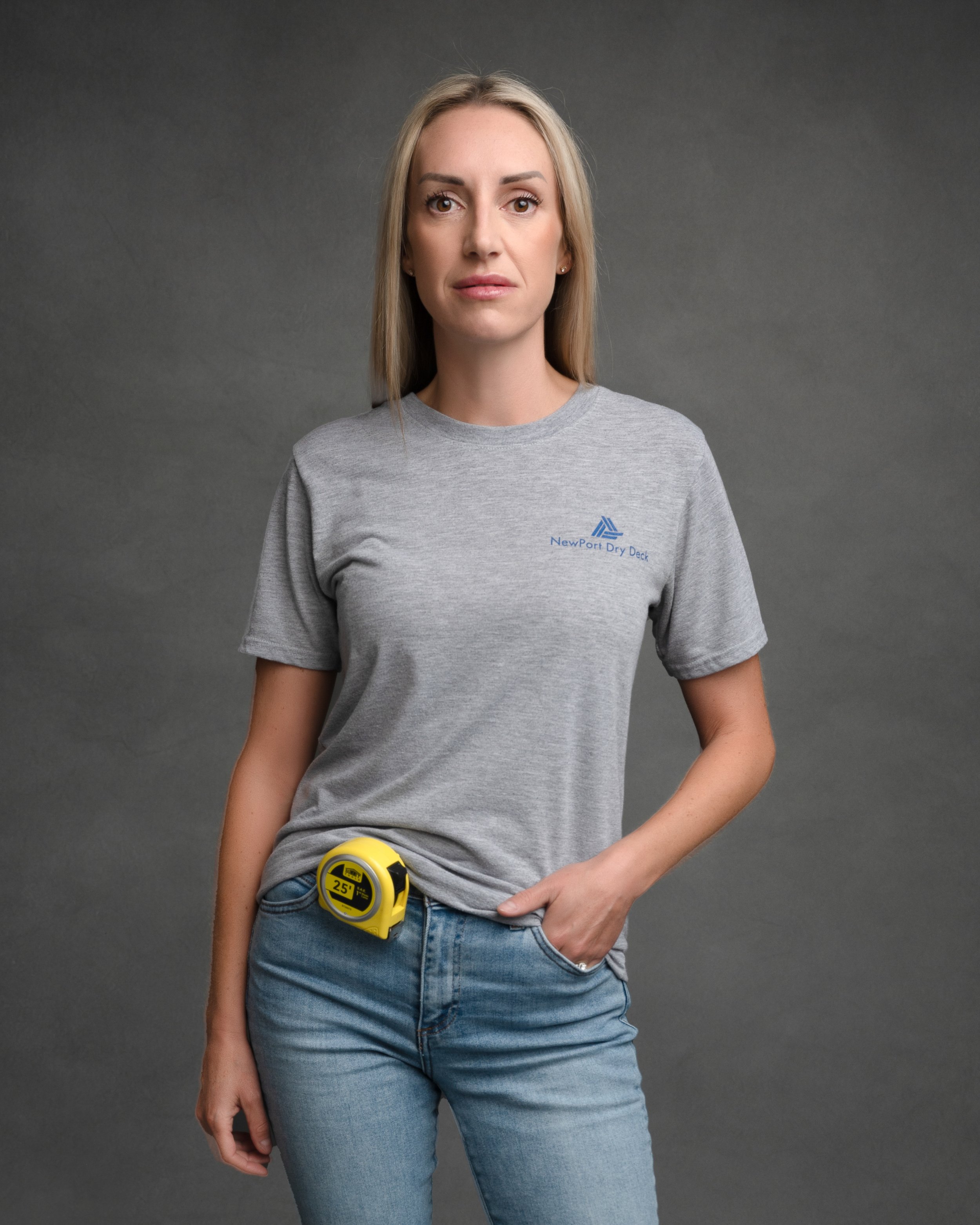 A woman with long blonde hair, wearing a gray NewPort Dry Deck t-shirt and blue jeans, standing against a gray background with a yellow measuring tape hanging from her waistband.