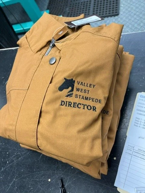 A folded tan uniform with a Valley West Stampede logo and the word 'DIRECTOR' embroidered on it, placed on a black surface.