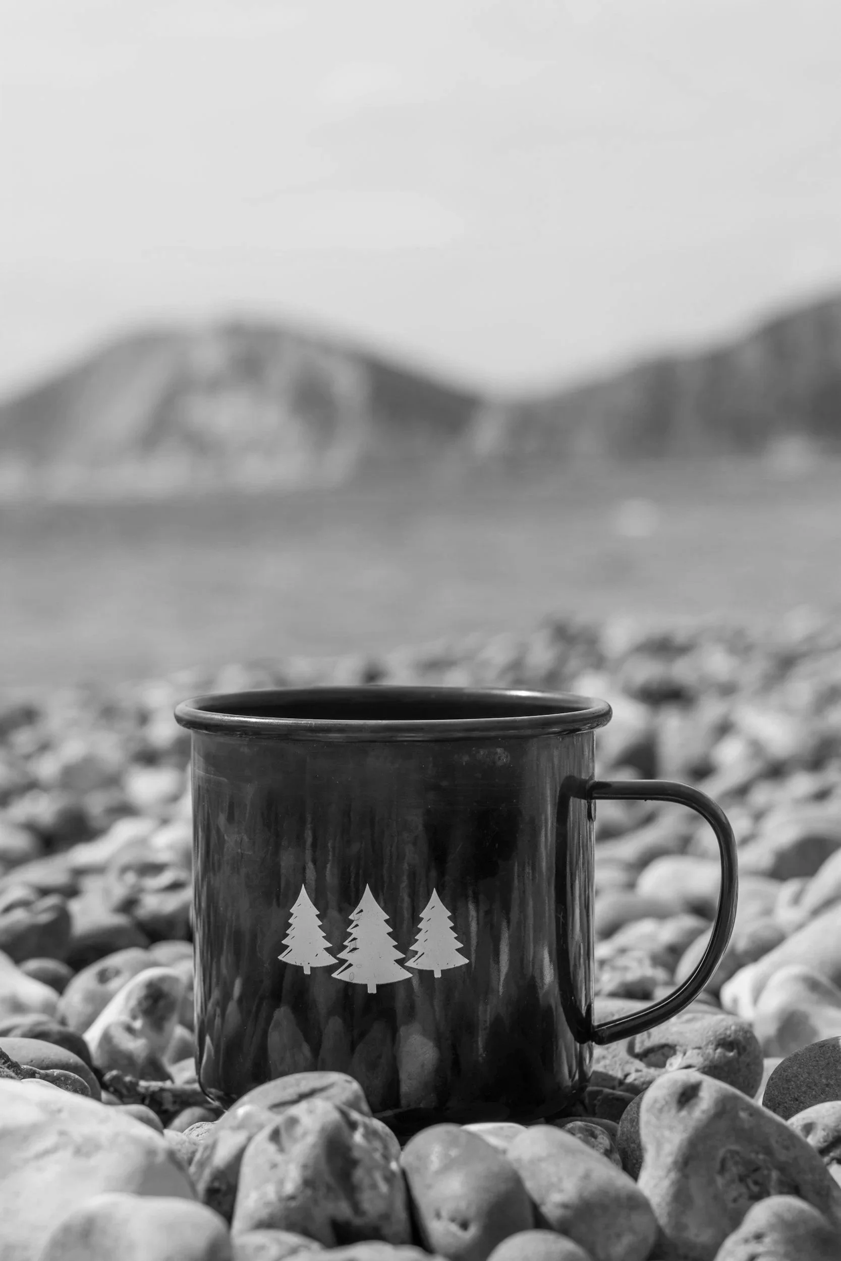 A black mug with Christmas tree silhouettes on its surface, sitting on a rocky shore with mountains in the distance and a cloudy sky above.