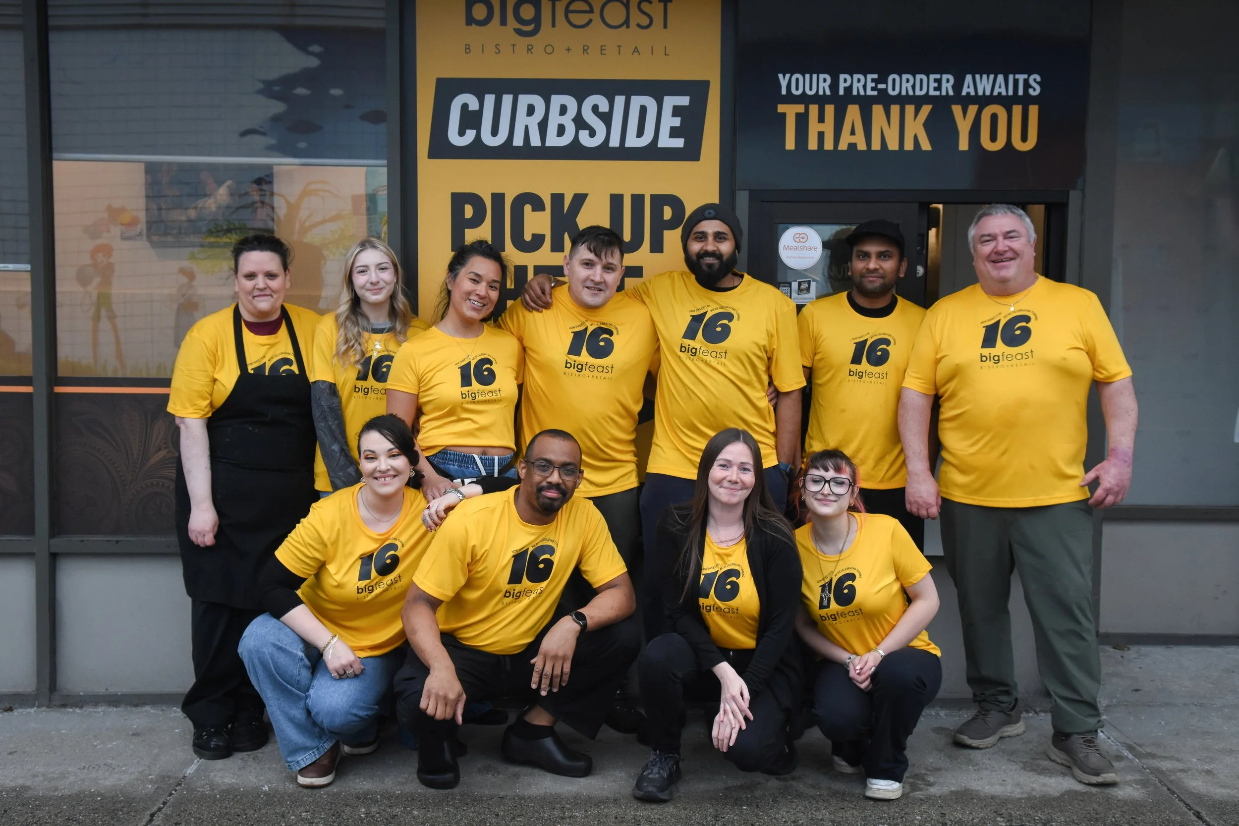Group of ten diverse people posing for a photo outside a restaurant, all wearing matching yellow T-shirts with the number 16 and the logo 'bigfeast,' some sitting and some standing in front of a large yellow and black sign that reads 'BIGFeast Bistro