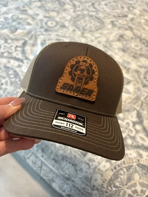 A brown and gray Richardson 112 trucker hat with a patch featuring a skull and mechanical gear logo.