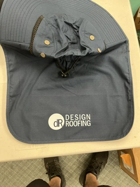 A round gray object with a hood and snap buttons, labeled 'Design Roofing,' placed on a table. A person's legs and feet are visible in the foreground.