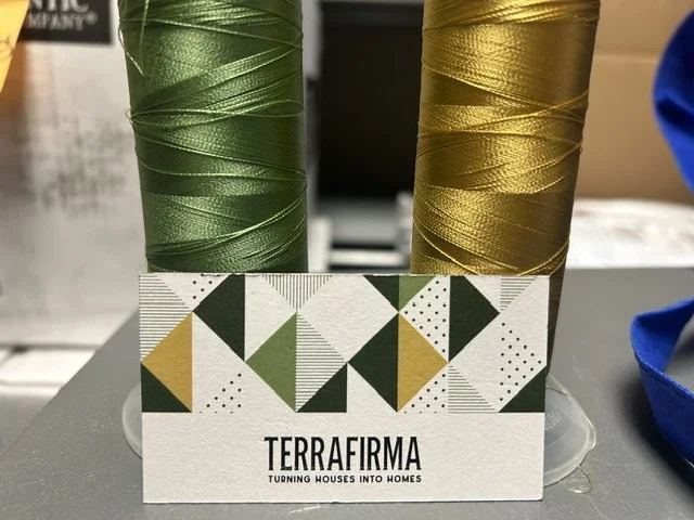 Two spools of metallic thread, one green and one gold, placed behind a business card with a geometric pattern and the logo 'Terrafirμα' with the tagline 'Turning houses into homes'.