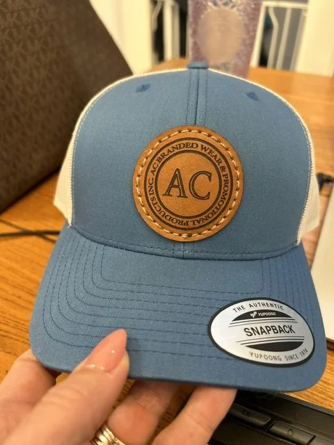 Blue and white snapback cap with a leather patch on the front reading 'AC' and encircled by the words 'Since a branded wear authorized production'.