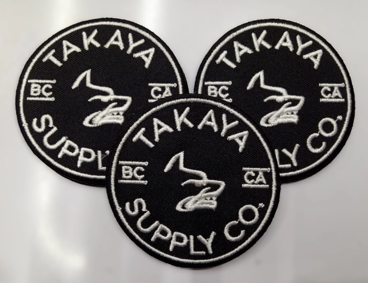 Three black circular patches with white embroidered text and logo. The patches display the words 'TAKAYA SUPPLY CO.', 'BC', and the number '2' with an outline of a bear's head.
