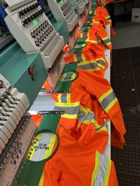 Multiple embroidered orange safety vests with reflective yellow stripes draped over sewing machines in a factory setting.