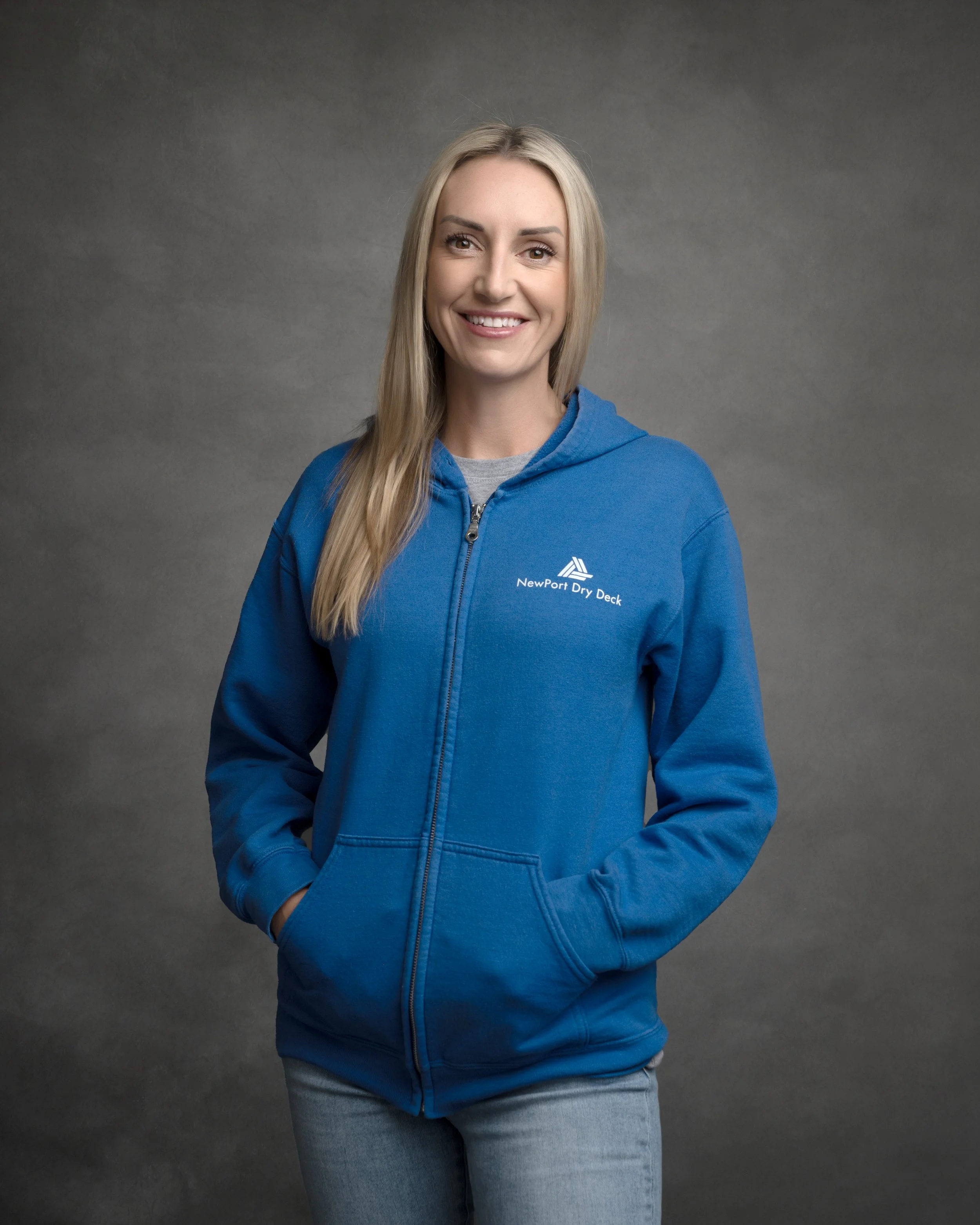 A woman with long blonde hair wearing a blue zip-up hoodie with a logo and gray jeans, standing against a gray background with a pleasant expression.