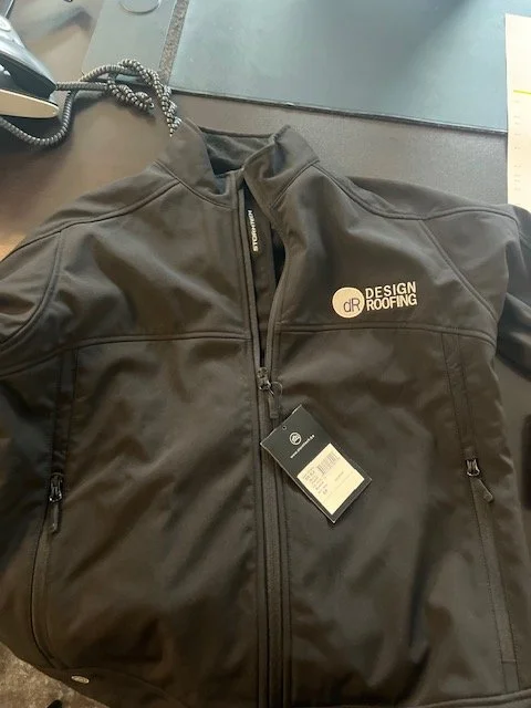 Black jacket with a zipper, logo that says 'Design Roofing,' and two zippered side pockets, placed on a desk with a phone and papers nearby.
