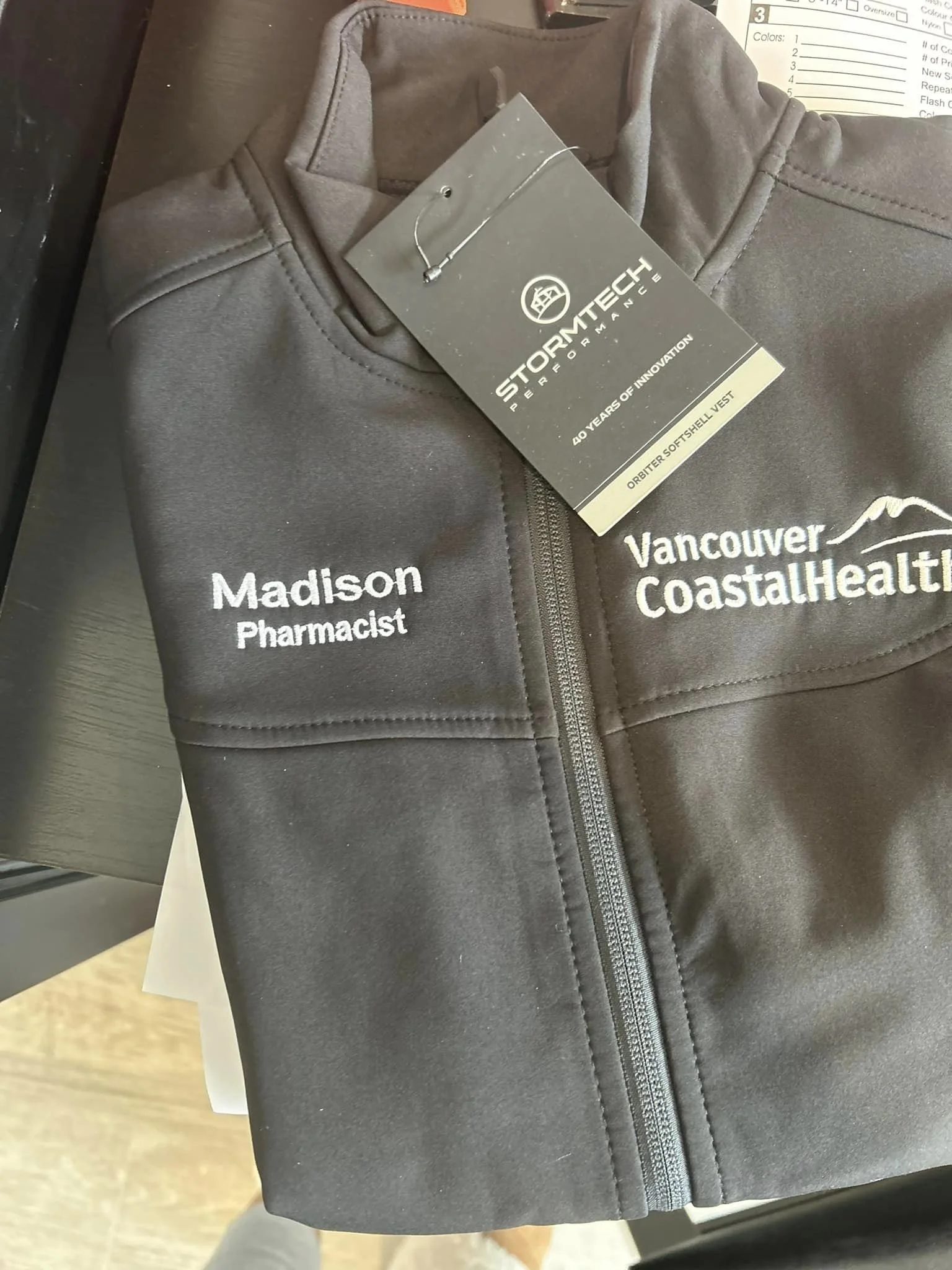 Close-up of a gray softshell vest with embroidered white text saying "Madison Pharmacist" on the left side and "Vancouver Coastal Health" on the right side, zippers and a black tag attached to the collar with the StoreMtech logo and text.