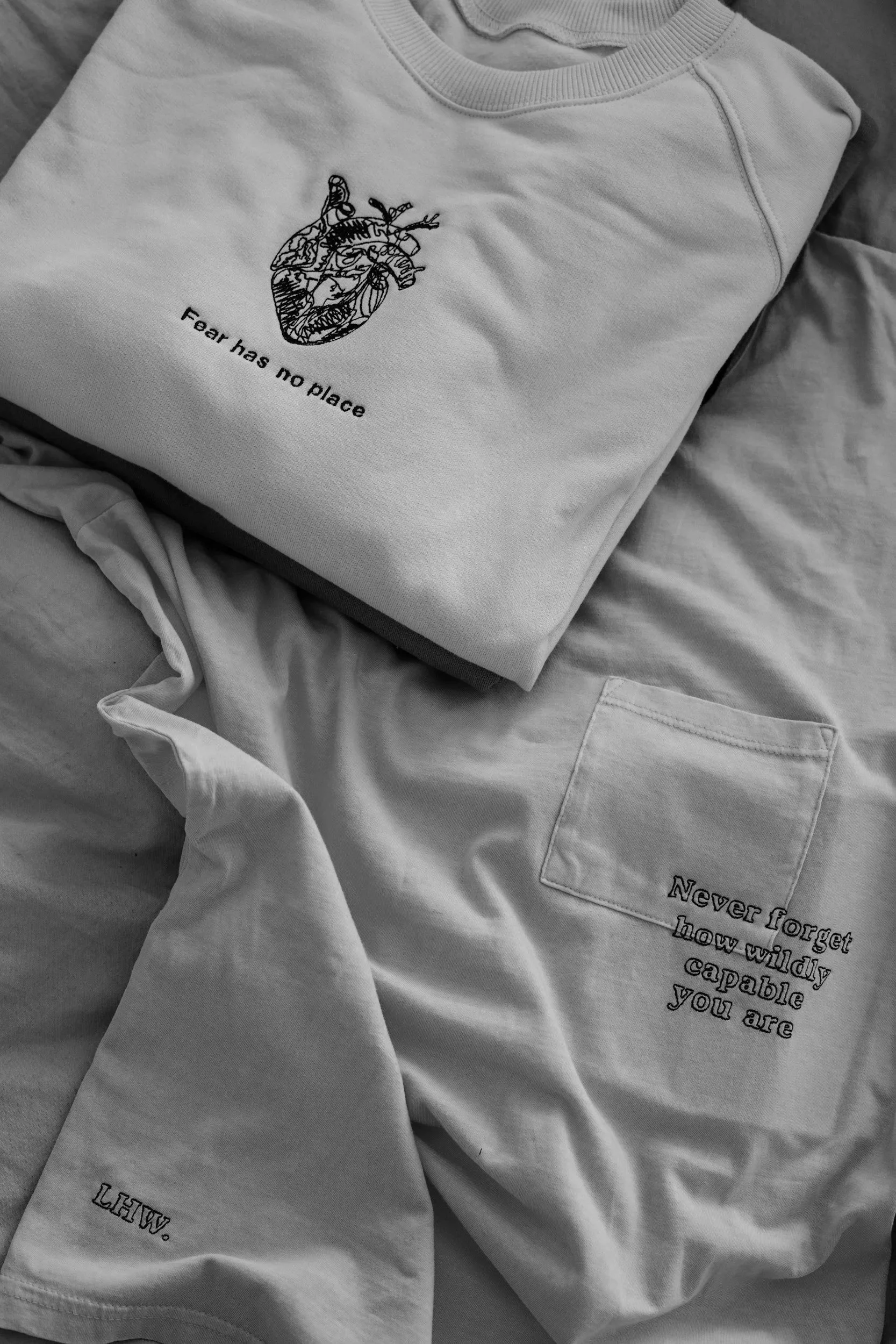 Gray sweatshirt with a line drawing of a human heart and the words "Fear has no place" below it, and part of a light-colored shirt with text that reads "Never forget how wildly capable you are".