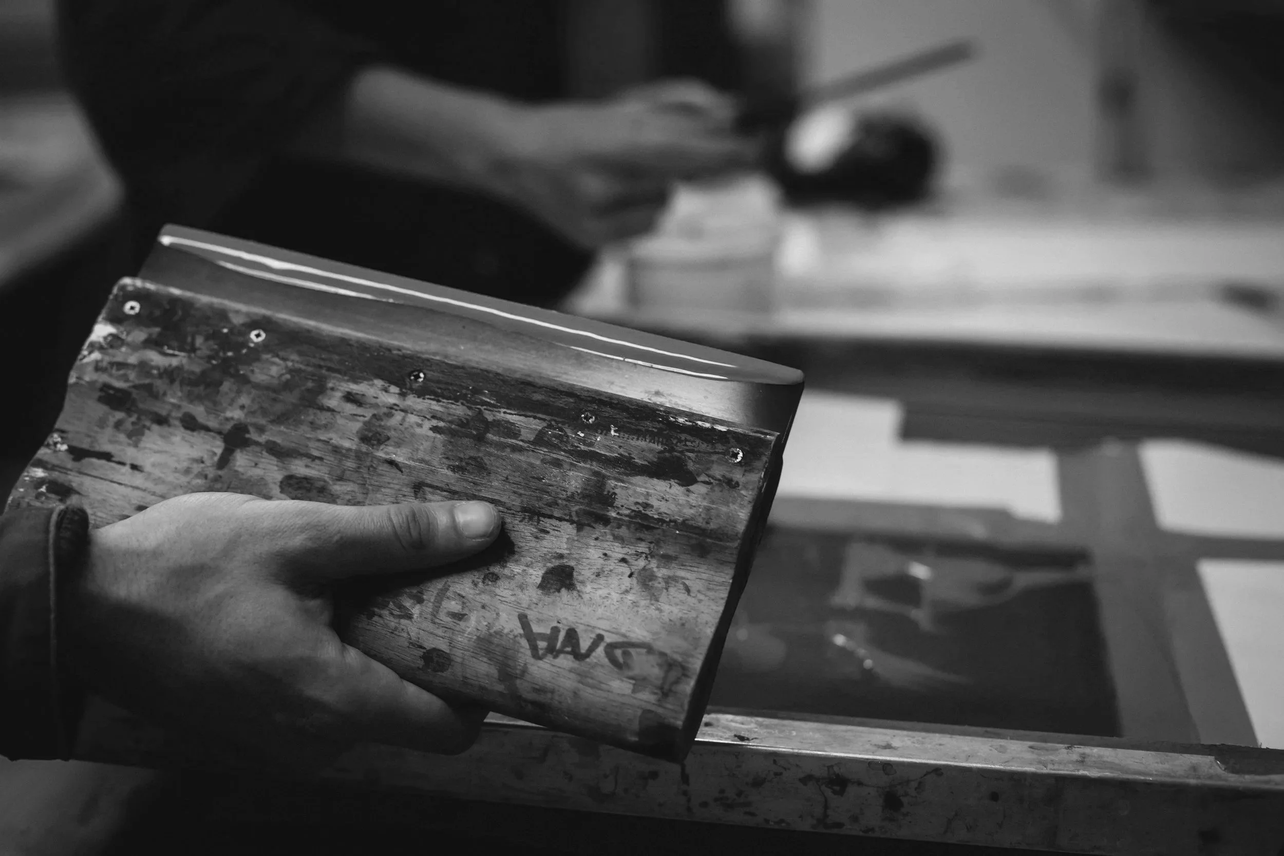 A person holding a weathered, paint-splattered wooden block in a workshop setting, with blurred tools and furniture in the background.