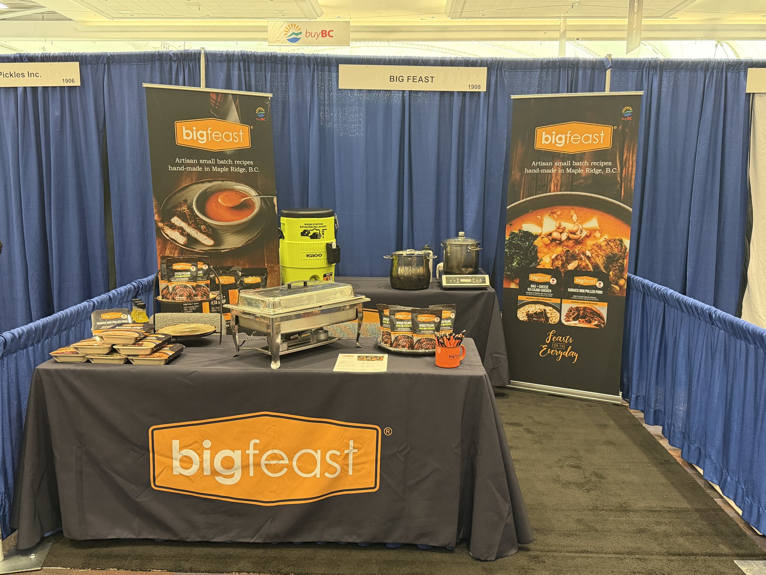 A booth at an event with blue curtains, promotional banners for Big Feast, and a table displaying packaged food products like baked goods and sauces, with containers and a wash station.