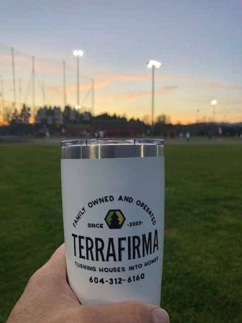 Hand holding a TeraFirma beverage can in front of a baseball field at dusk.