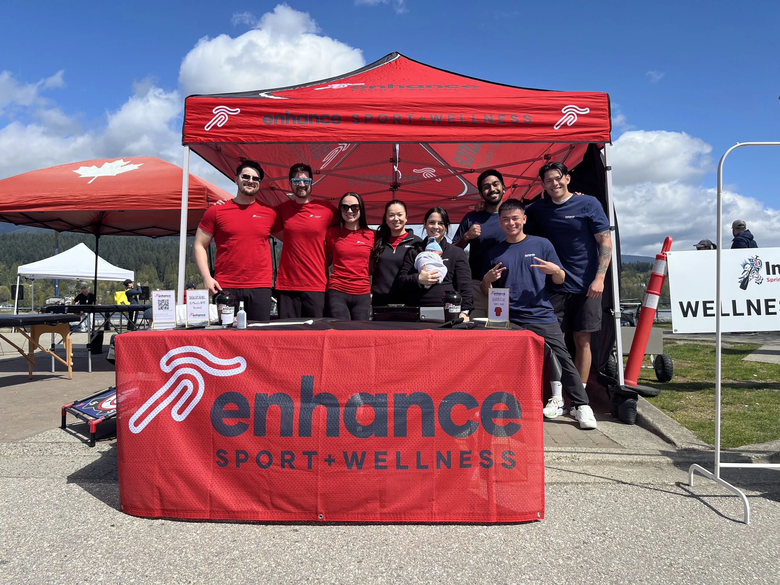Group of seven people standing behind a red table and under a red and black canopy, with a white and red logo that says 'enhance SPORT + WELLNESS,' outdoors on a sunny day with clouds in the sky.
