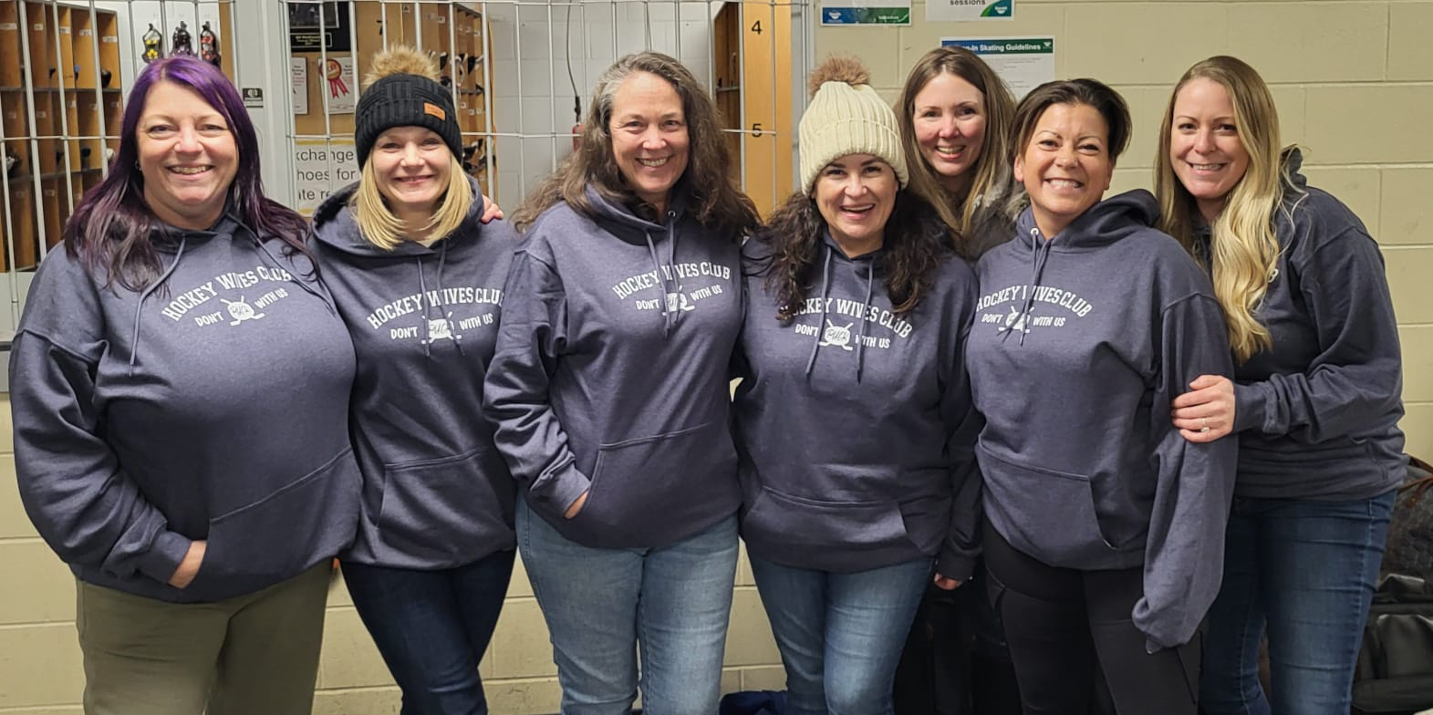 Group of seven women standing together in a hallway, all wearing matching dark gray hoodies with white text that reads 'Hockey Wives Club Don't F*** With Us'. They are smiling at the camera with lockers and a beige wall in the background.