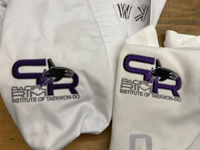White martial arts uniform with patches of the Pacific Rim Institute of Taekwondo, featuring the organization's logo with a purple 'PR' and a black and purple panther head.