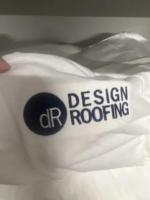 Close-up of a white fabric T-shirt with a logo and text that reads 'DR Design Roofing' in black.