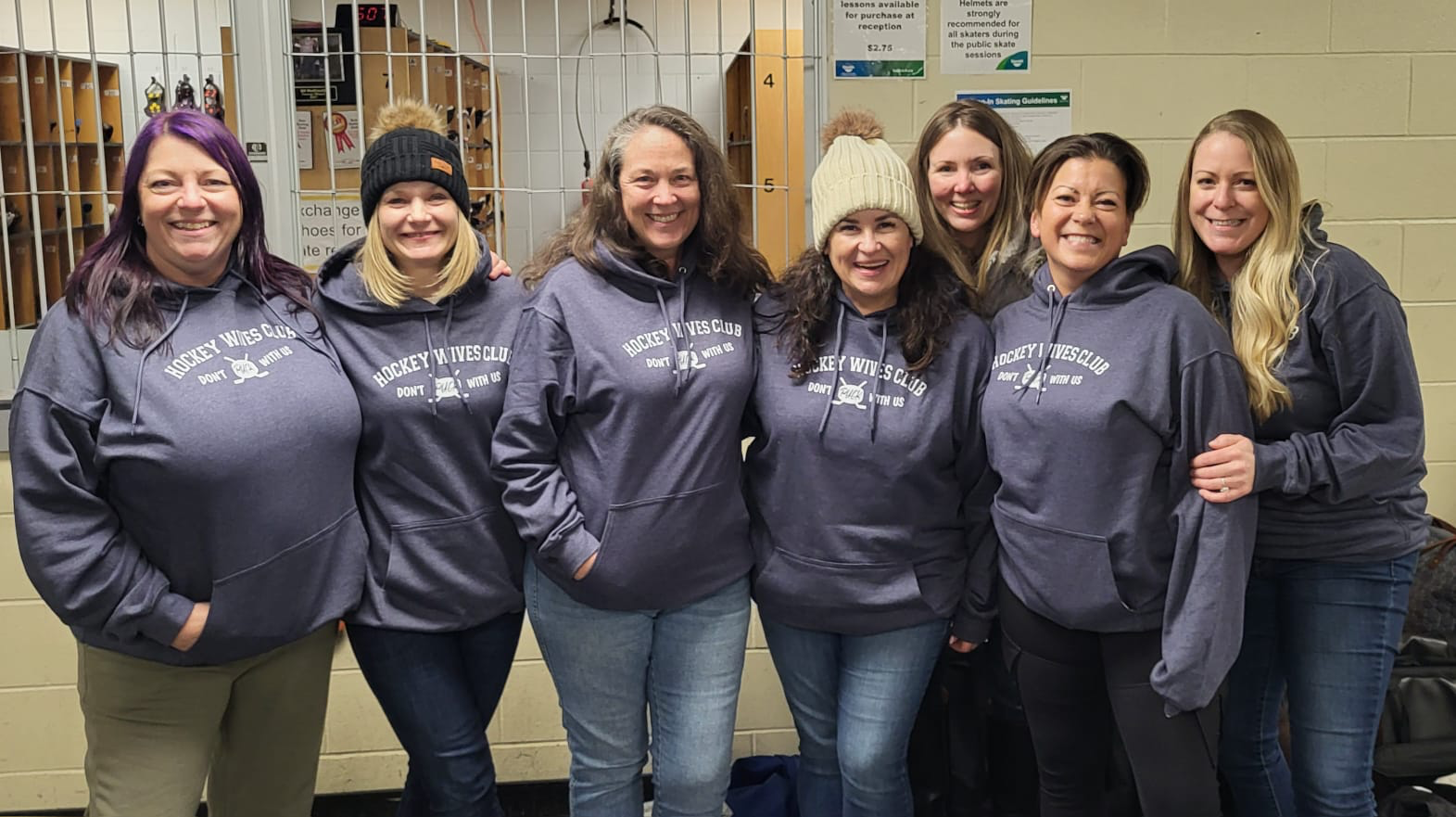 Group of eight women wearing gray hoodie sweatshirts with 'Hockey Wives Club' printed on them, standing together inside an ice skating rink locker room.