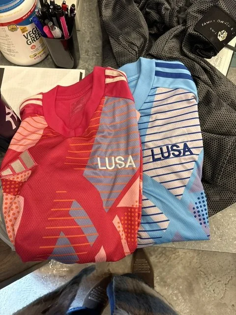 Two folded sports jerseys, one red and one blue, with 'LUSA' printed on the front; the red jersey has orange and pink accents, and the blue jersey has blue and white accents.