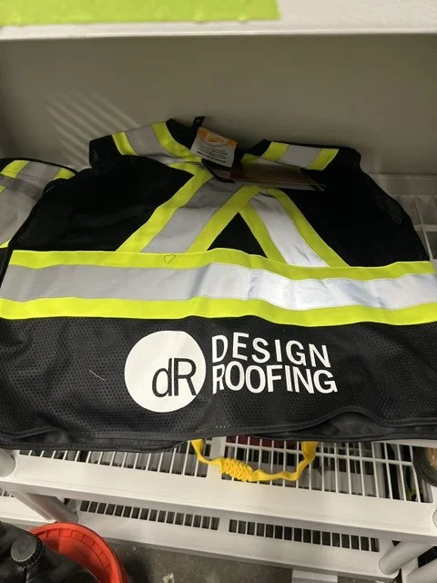High visibility safety vest with reflective strips and the logo for DR Design Roofing, placed on a white shelf.
