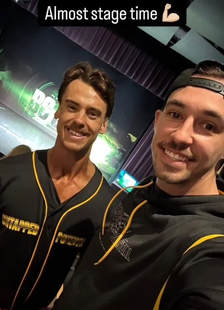 Two young men taking a selfie backstage at a gaming event, smiling, with stage lighting and screens in the background.