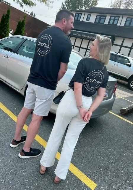 A man and woman wearing matching black T-shirts with a 'CityState' logo, standing in a parking lot near a silver car, engaged in conversation.