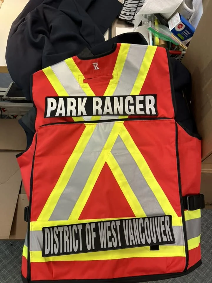 A red safety vest with yellow reflective stripes, labeled 'PARK RANGER' and 'DISTRICT OF WEST VANCOUVER'.