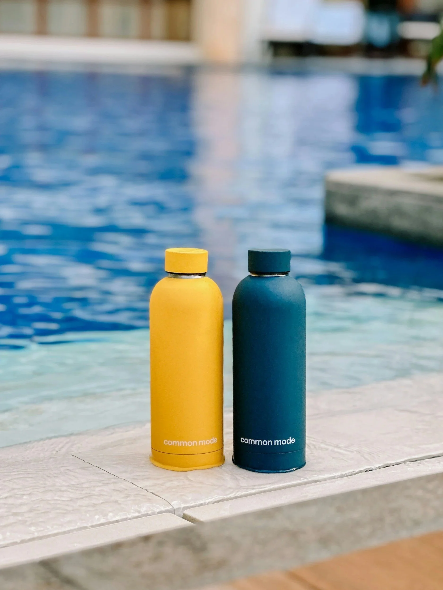Two cylindrical bottles, one yellow and one dark blue, labeled "common mode," are placed on a white surface near a swimming pool with blue water and a wooden deck.