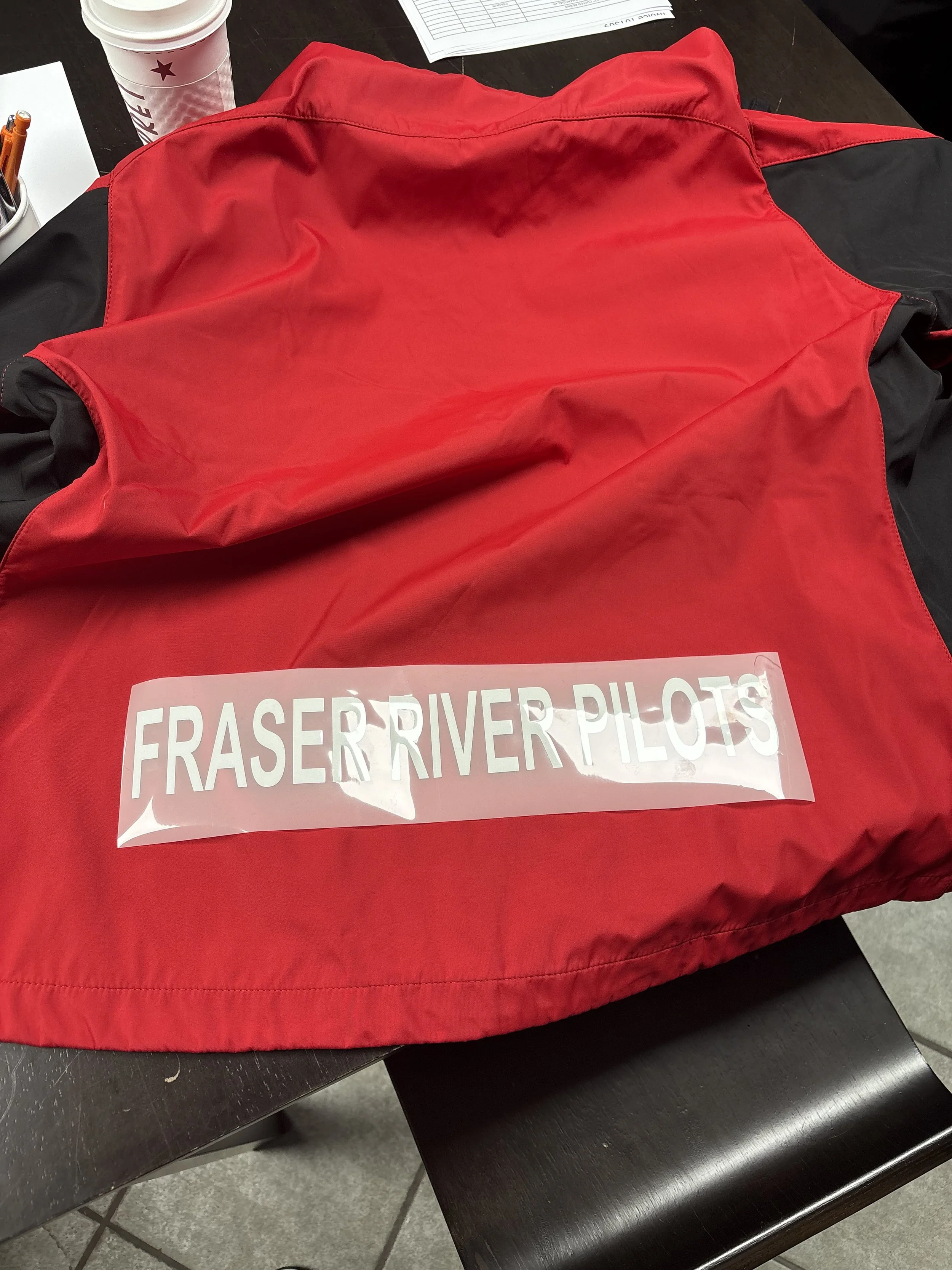 Red and black jacket with a white sign on top that reads 'FRASER RIVER PILOTS' placed on the front, on a dark table. In the background, a disposable cup with a lid and straw, and some papers are visible.