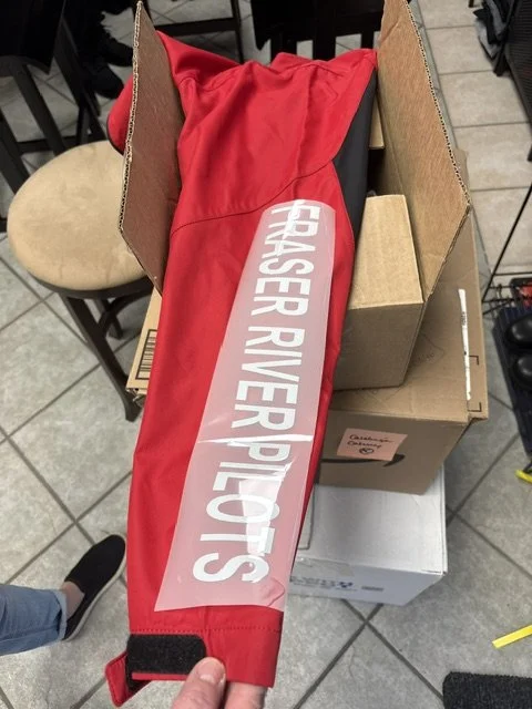 A red fabric item with a white label reading 'CRASER RIVER PILOTS' is placed inside an open cardboard box, on a tiled floor.