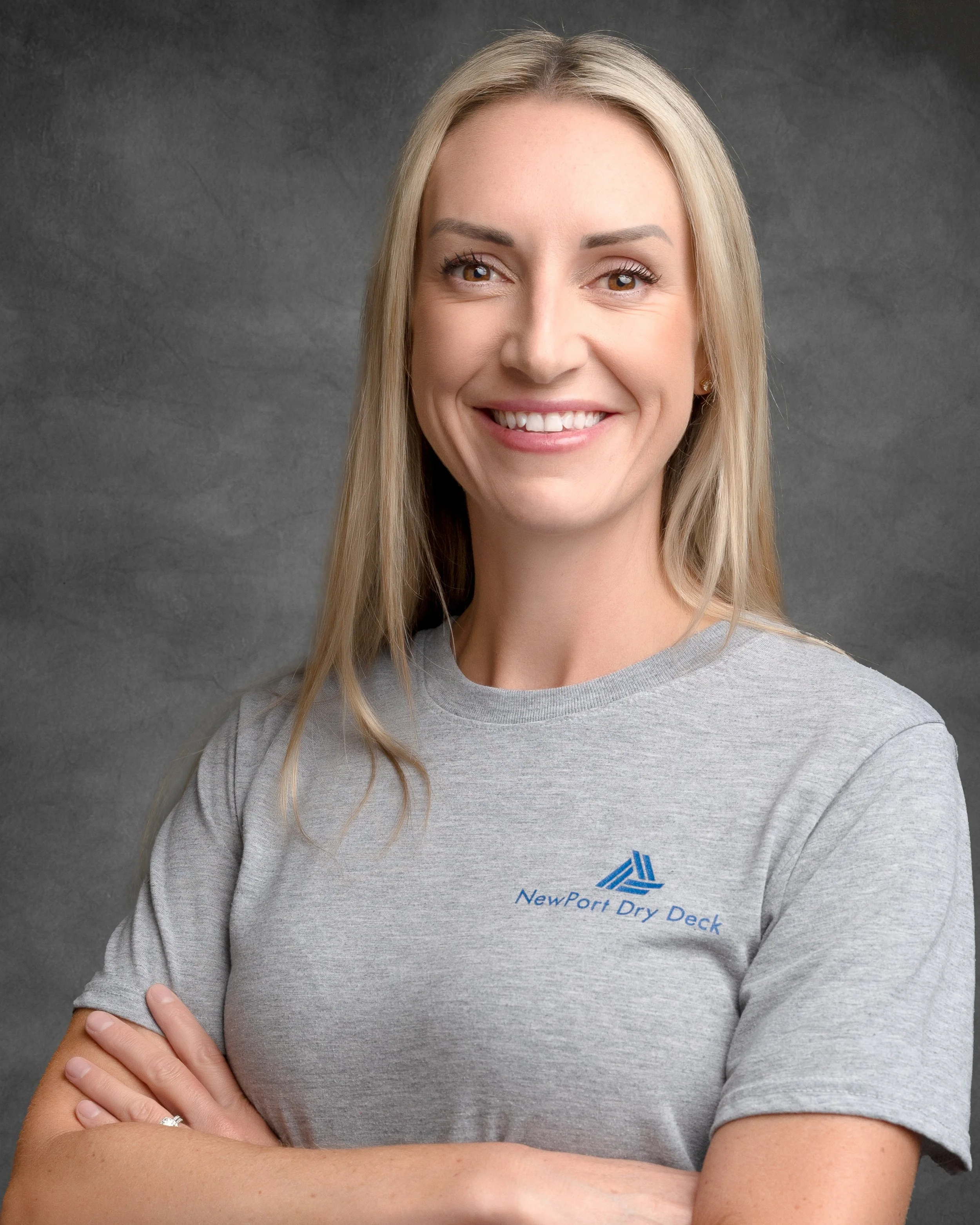A woman with long blonde hair and light skin, smiling and wearing a gray t-shirt with a company logo, crossing her arms against a gray background.