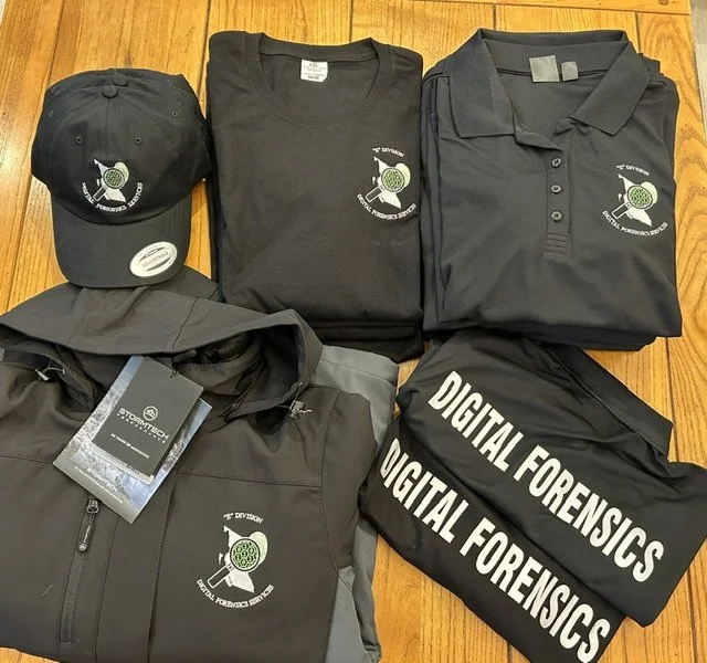 Collection of black and gray clothing and accessories with a forensic club logo, including a cap, two polo shirts, a jacket, and folded T-shirts with 'Digital Forensics' printed on them.