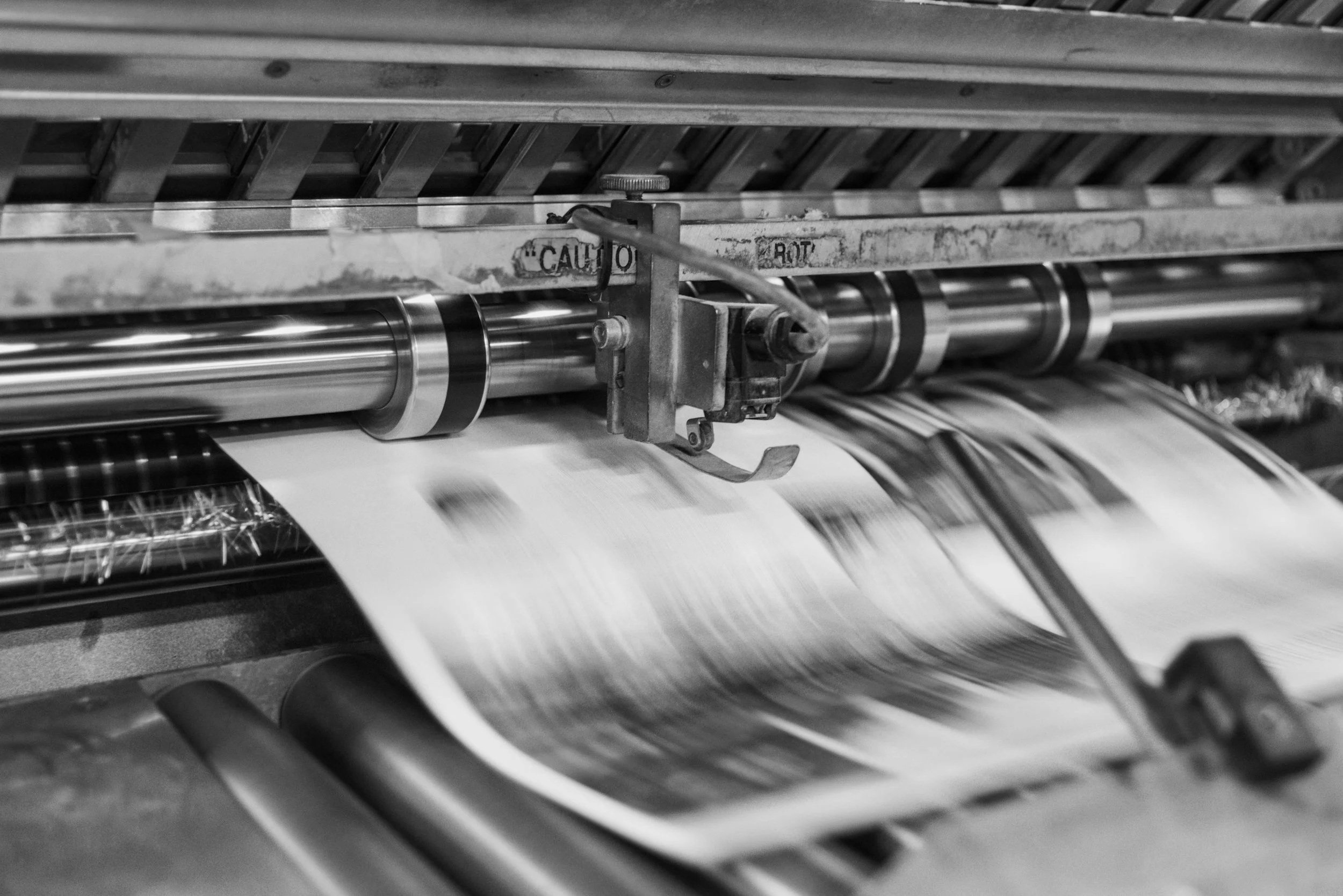 Close-up of a printing press with metal rollers and mechanisms, shown in black and white.