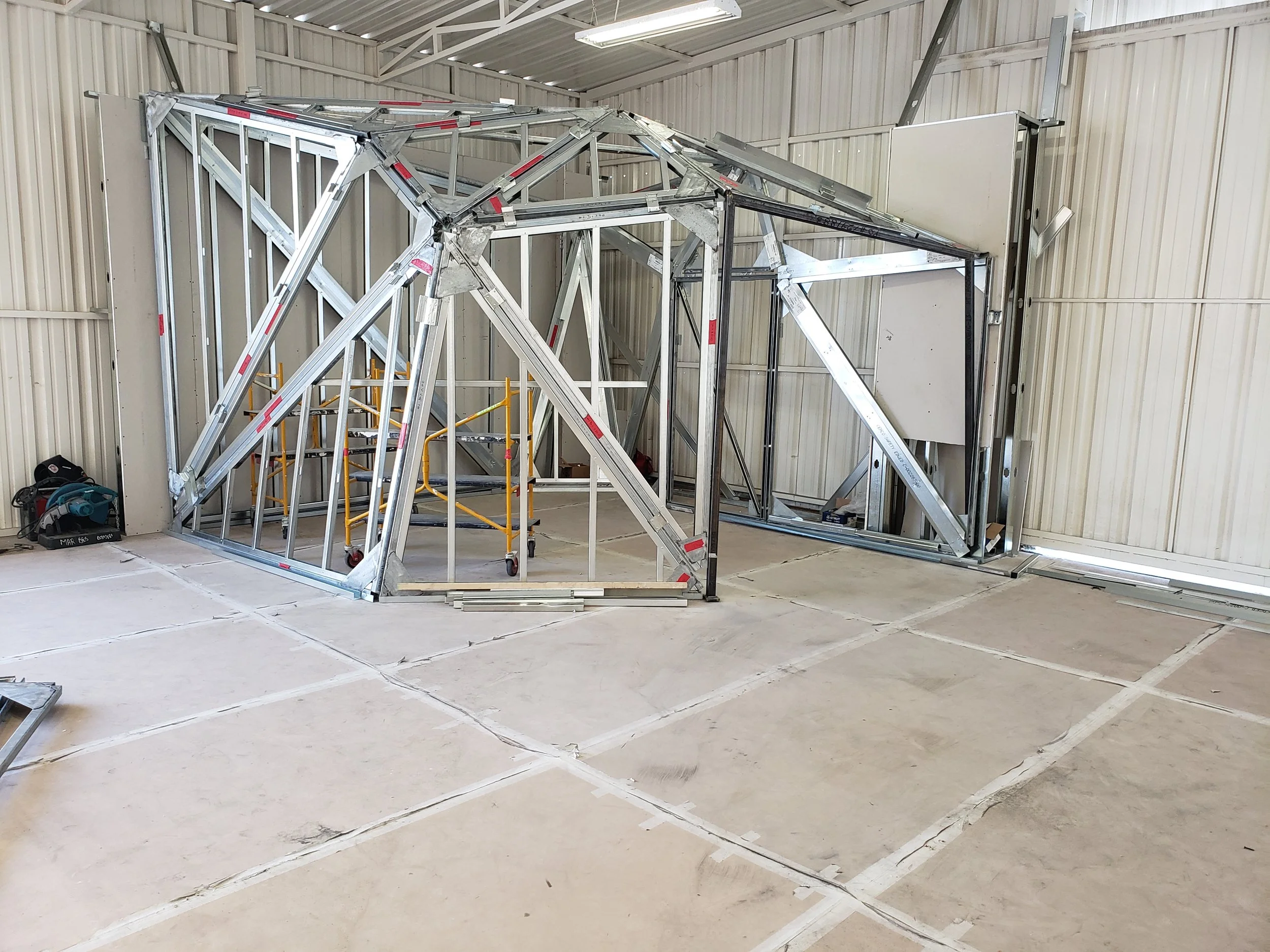 Construction site with metal framing for a structure inside a workshop with concrete floor and metal walls.