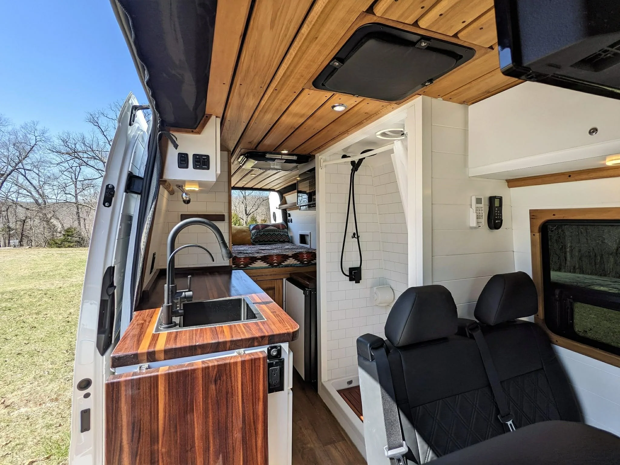 Interior of a converted camper van with a small kitchen area featuring a wooden countertop, a sink, and cabinets. There is a bathroom with a shower visible behind a white curtain, and a bed area with cushions at the rear of the vehicle. Black seats with seat belts are also visible.