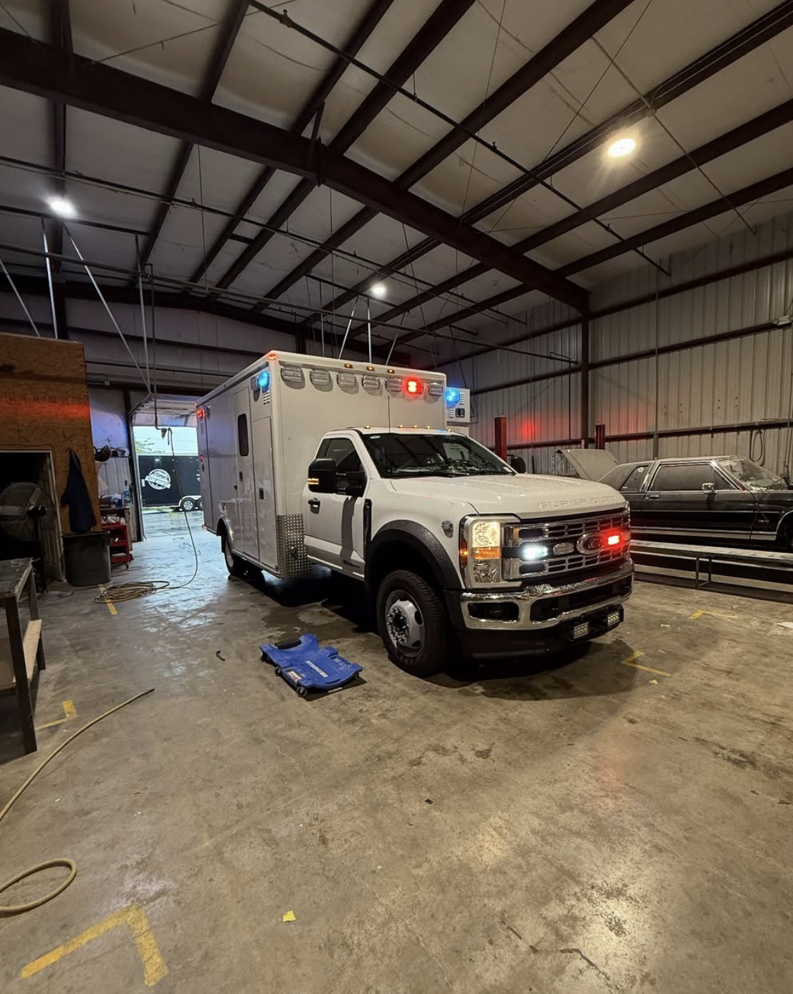 Type 1 ambulance inside a garage with its lights on, next to a black classic car, and various tools and equipment around.