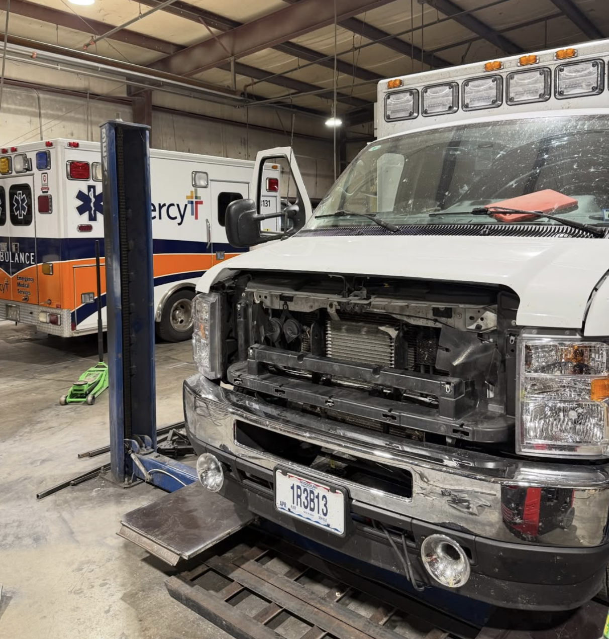 An ambulance and a damaged white emergency vehicle inside a garage or repair shop, with the front of the damaged vehicle missing its grille and components.