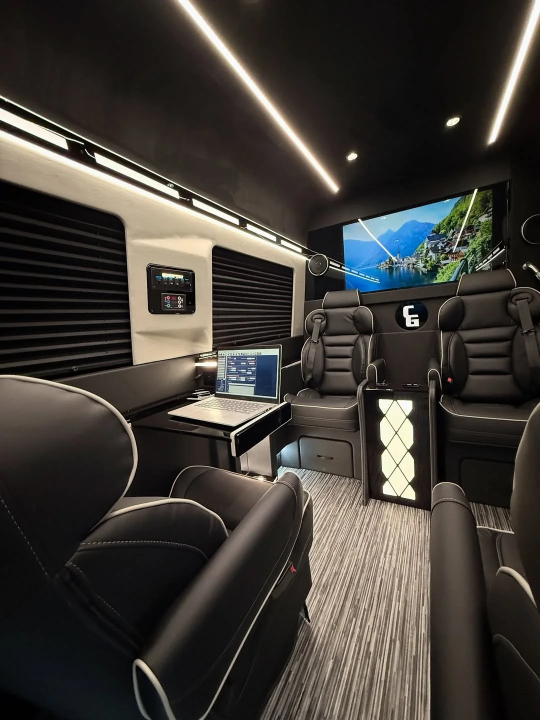 Jet style Luxury interior of a modern vehicle or mobile office with black leather seats, a computer on a small table, a large TV screen displaying a scenic landscape, and ambient lighting.