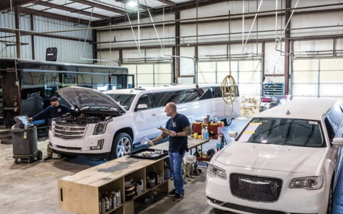 Two mechanics working on white vehicles inside a garage or workshop with tools and equipment around.