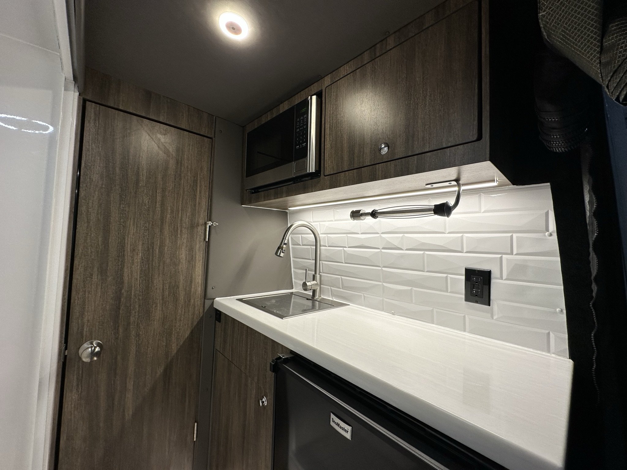 Small kitchen with dark wood cabinets, microwave, white backsplash, stainless steel faucet, small countertop, and mini fridge in a Mercedes Sprinter